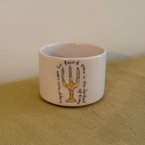 May include: A small, off-white ceramic candle holder with a gold-colored menorah design. The holder has handwritten text that reads "happiness can be found even in the darkest of times."