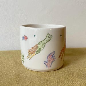 May include: White ceramic mug featuring hand-drawn illustrations of a mermaid, fish, and coral in various colours. The mermaid has green scales and brown hair. The mug is cylindrical with a smooth finish.
