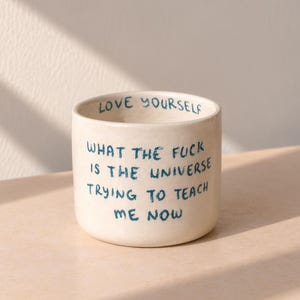 Może przedstawiać: Cylindryczny kubek ceramiczny w kolorze złamanej bieli z ciemnoniebieskim, ręcznie malowanym tekstem. Wewnętrzna krawędź zawiera napis LOVE YOURSELF, a na zewnątrz widnieje tekst WHAT THE FUCK IS THE UNIVERSE TRYING TO TEACH ME NOW pisany odręcznie.