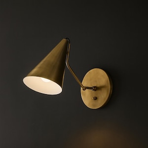 May include: A brass wall light with a conical lampshade and a circular backplate. The sconce is fixed to a dark wall, and the light from the bulb shines downwards. The design is minimalist and modern, suitable for bedroom or living room lighting.