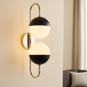 Könnte beinhalten: Moderne Wandleuchte mit zwei kugelförmigen Leuchten. Jede Kugel ist halb schwarz und halb weiß, mit einem polierten goldfarbenen Metallrahmen. Die Leuchte ist an einer beigefarbenen Wand montiert.