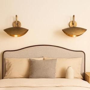 Könnte beinhalten: Zwei Wandleuchten aus Messing mit geschwungenem Design sind über einem Bett montiert. Die Leuchten haben einen warmen, goldenen Farbton und in jeder ist eine Glühbirne sichtbar. Das Bett hat ein helles Kopfteil, Kissen und eine dekorative Vase.