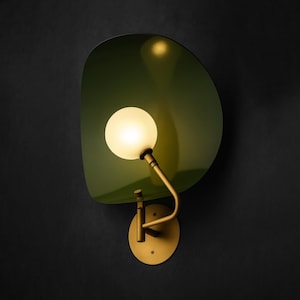 May include: A wall-mounted lamp with a green, leaf-shaped shade and a spherical, glowing bulb. The lamp has a brass-colored arm and a circular base, mounted on a dark background. The design is modern and stylish.