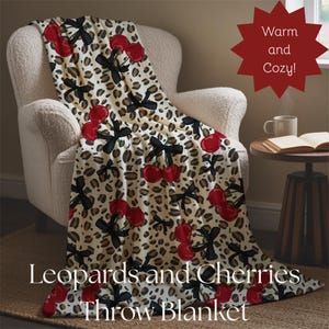 May include: A throw blanket with a cream background featuring a leopard print pattern, red cherries, and black bows. The blanket is draped over a white armchair. The text "Leopards and Cherries Throw Blanket" is at the bottom.