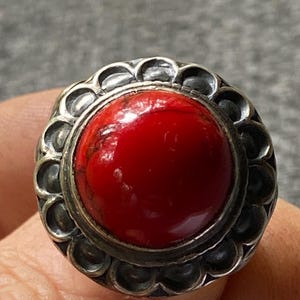 Könnte beinhalten: Ein Ring mit einem großen, runden, polierten roten Stein in einer silbernen Fassung. Die Fassung ist mit einem gewellten, blütenblattartigen Rand verziert, der den zentralen Stein umschließt und einen strukturierten Rahmen um die rote Fläche bildet.