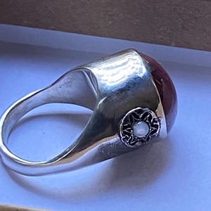 May include: A polished silver ring featuring a large, smooth, dark red cabochon stone set in the center. The side of the wide silver band is decorated with a small, circular, ornate metal emblem containing a tiny, iridescent white pearl.