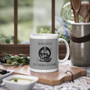May include: White ceramic mug with a gray band featuring a black skull design, a candle, and the text "ROMANCE BUT MAKE IT DARK." The mug is on a wooden cutting board with olives and a container of utensils in the background.