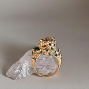 May include: A gold ring with a jaguar design. The jaguar's head is decorated with green and white gemstones, and a red gemstone eye. The ring is displayed on a clear, crystalline surface.