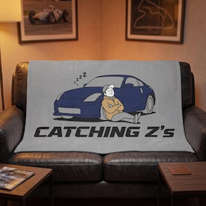 May include: A light gray fleece blanket featuring a graphic of a person sleeping against a dark blue sports car. The design includes the text CATCHING Z'S in a bold black font, with small black Z letters floating above the person's head.