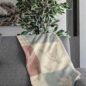 May include: A soft throw blanket with a patchwork design featuring dove illustrations and floral accents. The blanket has a color palette of cream, dusty rose, and light blue, draped over a grey sofa. The blanket is approximately 127 x 152 cm.