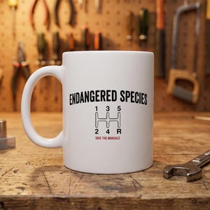 May include: A white ceramic mug featuring the black text ENDANGERED SPECIES above a black manual transmission gear shift pattern diagram with numbers 1, 2, 3, 4, 5, and R, and the red text SAVE THE MANUALS printed below the gear pattern.