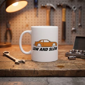 Op de afbeelding: Een witte keramische mok met een grafische illustratie van een oranje coupé auto in profiel. Onder de auto staat de tekst "LOW AND SLOW" gedrukt in vette, zwarte, schreefloze hoofdletters. De mok heeft een standaard handvat en cilindrische vorm.