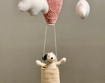 May include: A pink knitted hot air balloon with a white knitted cloud and a white knitted moon hanging from a white felt ring. A knitted dog is in the basket of the balloon.