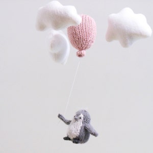 Pu&ograve; includere: Un mobile di pinguino lavorato a maglia con un palloncino rosa, nuvole bianche e dettagli grigi e bianchi.