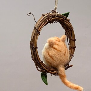May include: A felted orange and white cat figurine hanging from a twig wreath. The cat is facing away from the viewer and has a long tail.