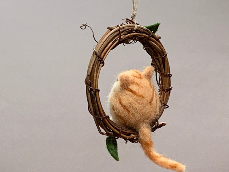 May include: A felted orange and white cat figurine hanging from a twig wreath. The cat is facing away from the viewer and has a long tail.