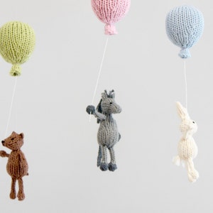 Pu&ograve; includere: Un mobile per neonati in maglia con un orso marrone, un cavallo grigio, un coniglio bianco e tre palloncini in verde, rosa e blu.