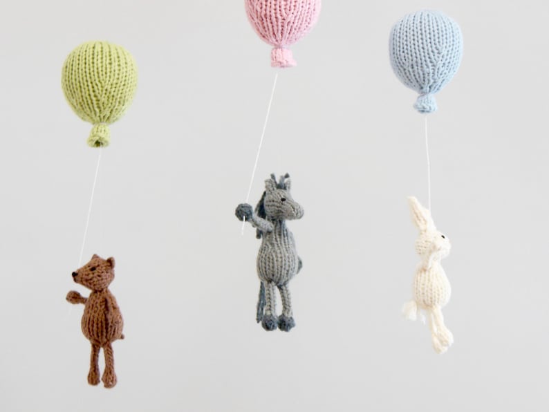 Pu&ograve; includere: Un mobile per neonati in maglia con un orso marrone, un cavallo grigio, un coniglio bianco e tre palloncini in verde, rosa e blu.