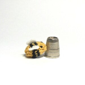 May include: A small crocheted bumble bee with yellow and black stripes and white wings, next to a silver thimble.