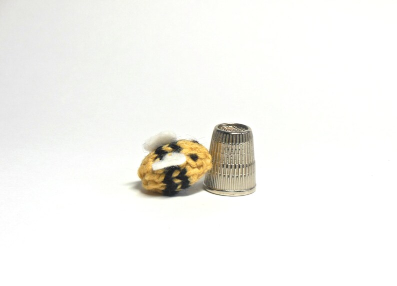 May include: A small crocheted bumble bee with yellow and black stripes and white wings, next to a silver thimble.