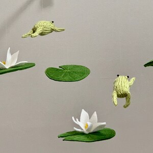 May include: A green felt frog mobile with white felt lily pads and flowers hanging from a wooden branch.