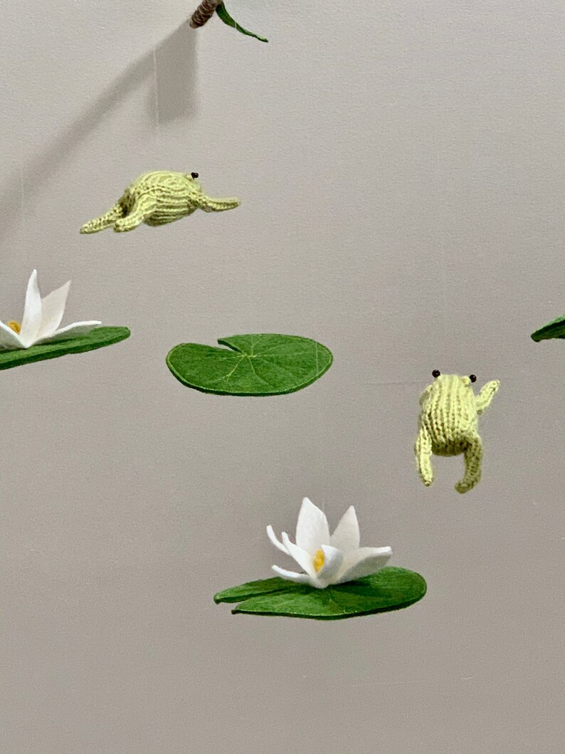 May include: A green felt frog mobile with white felt lily pads and flowers hanging from a wooden branch.