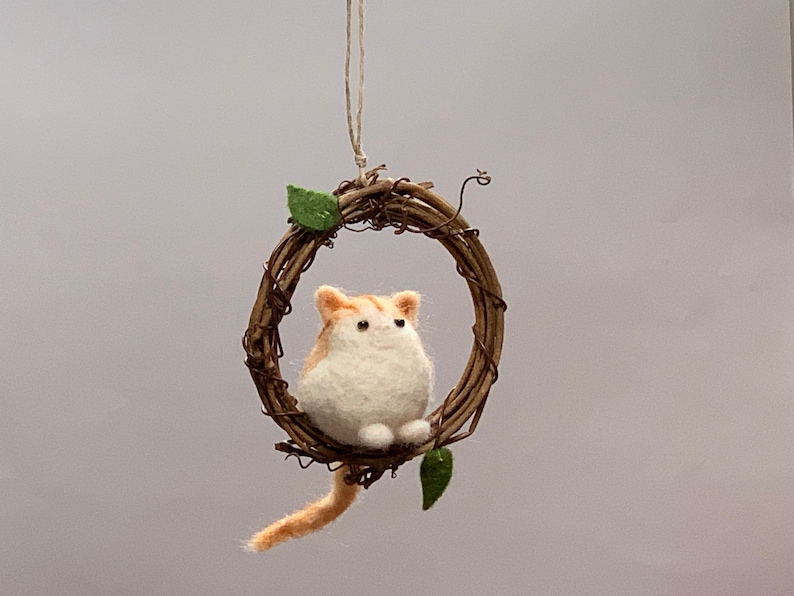 May include: A felted orange and white cat ornament hanging from a twig wreath with green leaves.