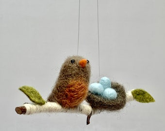 May include: A felt robin perched on a twig with a nest of blue eggs. The robin is brown with an orange breast. The nest is made of brown felt and the eggs are light blue.