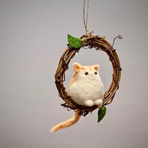 May include: A small, white and orange felted cat figurine sitting in a brown twig wreath with green leaves. The wreath is hanging from a string.