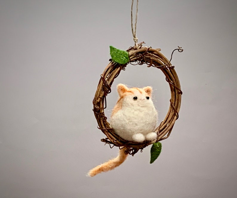 May include: A small, white and orange felted cat figurine sitting in a brown twig wreath with green leaves. The wreath is hanging from a string.