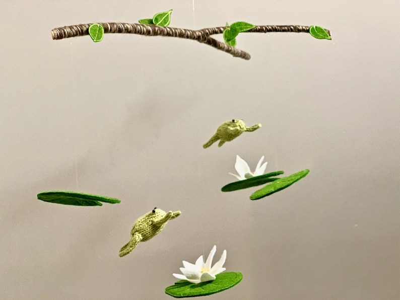 May include: A green frog mobile with three frogs hanging from a branch with green leaves. Each frog is hanging from a green felt lily pad. Two of the lily pads have white felt flowers on top.
