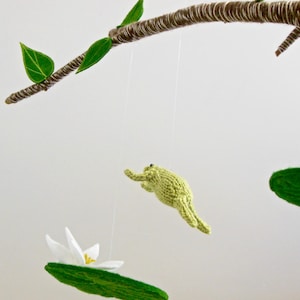 May include: A mobile with a green frog, a white flower, and green leaves hanging from a branch. The mobile is made of felt and yarn.