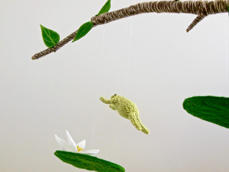 May include: A mobile with a green frog, a white flower, and green leaves hanging from a branch. The mobile is made of felt and yarn.