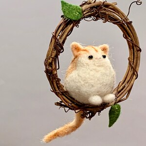 May include: A felted orange and white cat figurine hanging from a brown twig wreath with green leaves. The cat is sitting with its tail hanging down.