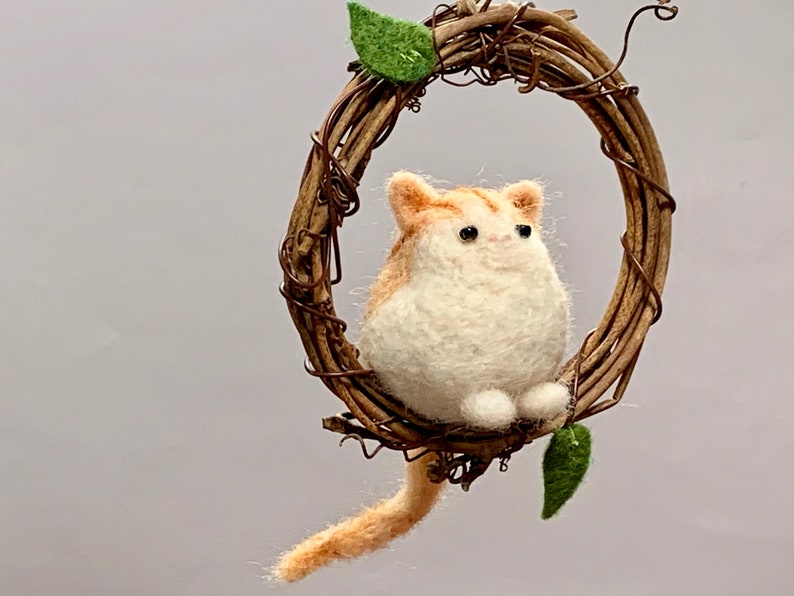May include: A felted orange and white cat figurine hanging from a brown twig wreath with green leaves. The cat is sitting with its tail hanging down.