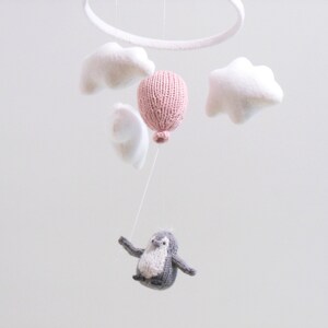 Pu&ograve; includere: Un mobile per bambini con un palloncino rosa lavorato a maglia, nuvole bianche in feltro e un pinguino grigio lavorato a maglia appeso a un anello bianco.