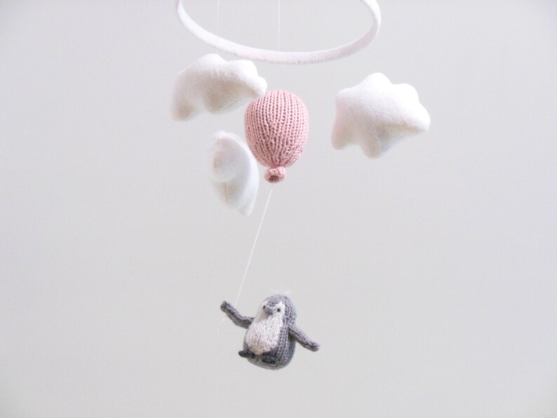 Pu&ograve; includere: Un mobile per bambini con un palloncino rosa lavorato a maglia, nuvole bianche in feltro e un pinguino grigio lavorato a maglia appeso a un anello bianco.