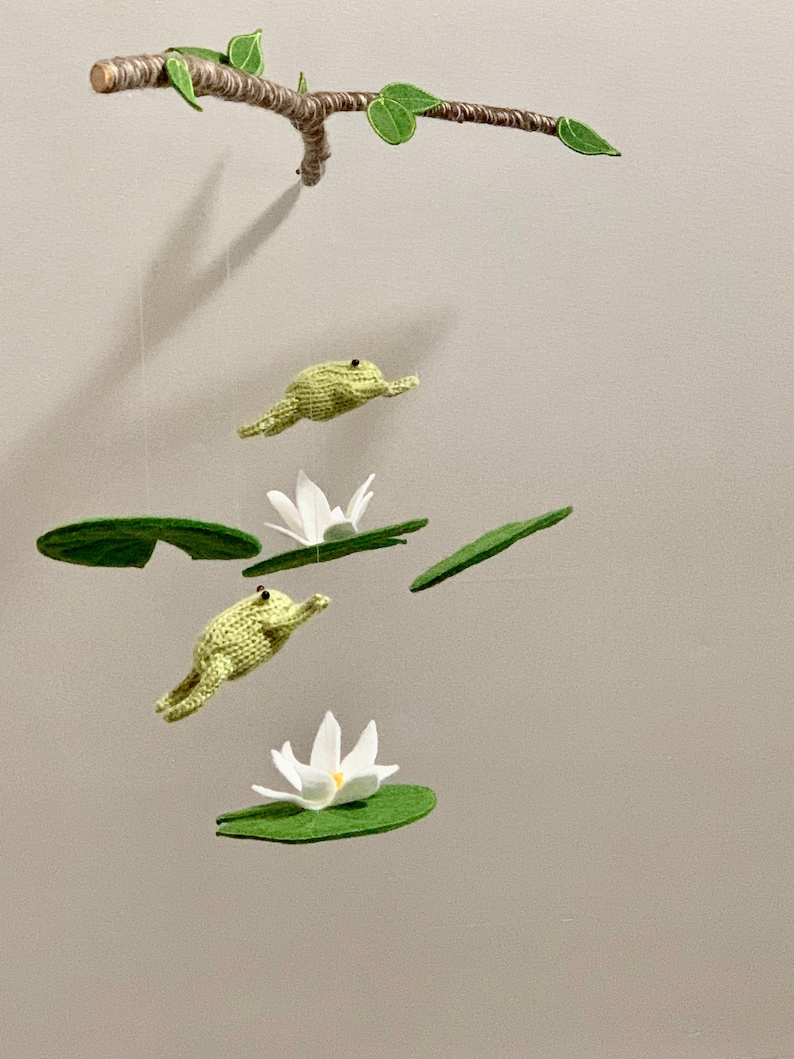 May include: A green felt mobile with three frogs and three lily pads. The frogs are jumping and the lily pads have white flowers.