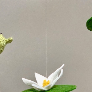 May include: A green crocheted frog hanging from the ceiling with a white crocheted lily pad and flower below it.