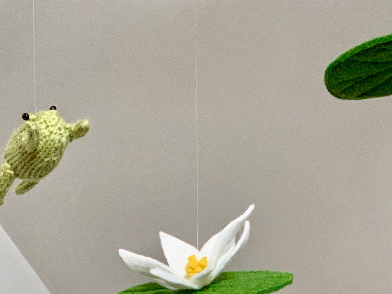 May include: A green crocheted frog hanging from the ceiling with a white crocheted lily pad and flower below it.
