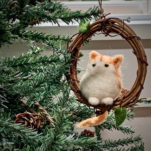 May include: A felted orange and white cat ornament hanging from a twig wreath with green leaves. The ornament is hanging from a Christmas tree.