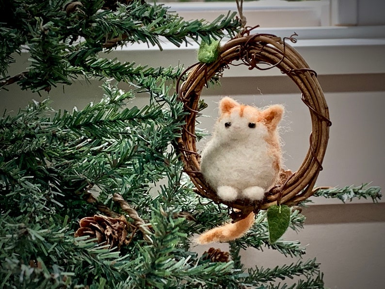 May include: A felted orange and white cat ornament hanging from a twig wreath with green leaves. The ornament is hanging from a Christmas tree.