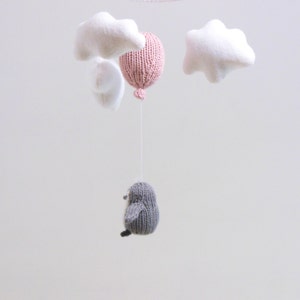 Pu&ograve; includere: Un mobile per neonati in maglia con un palloncino rosa, due nuvole bianche e un uccello grigio.