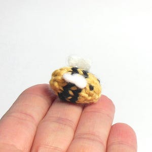May include: A small crocheted bee with black and yellow stripes and white wings. The bee is sitting on a person's hand.