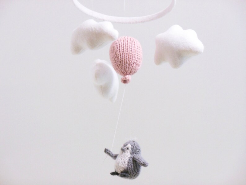 Pu&ograve; includere: Un mobile per neonati con un palloncino rosa lavorato a maglia, nuvole bianche e un pinguino grigio lavorato a maglia appeso a un anello di metallo bianco.
