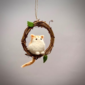 May include: A small, white and orange felted cat figurine sitting in a brown twig wreath with green leaves. The cat is sitting upright with its paws tucked under its body.
