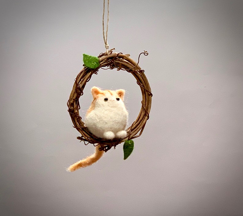 May include: A small, white and orange felted cat figurine sitting in a brown twig wreath with green leaves. The cat is sitting upright with its paws tucked under its body.