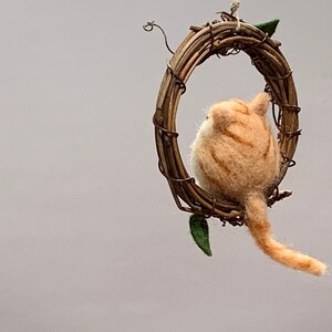 May include: A felted orange tabby cat figurine sitting inside a twig wreath. The cat is looking to the left, and its tail is hanging down.