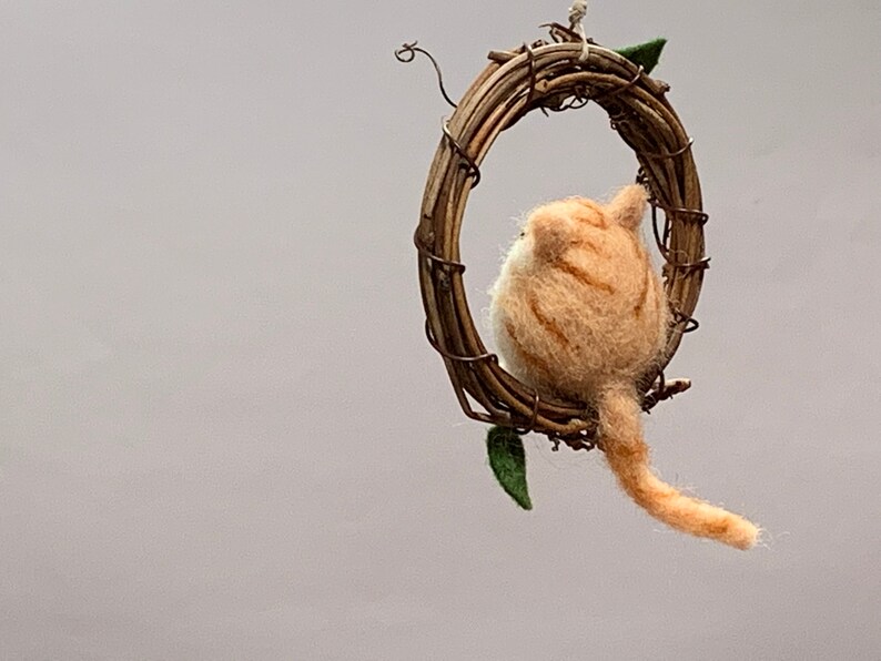 May include: A felted orange tabby cat figurine sitting inside a twig wreath. The cat is looking to the left, and its tail is hanging down.
