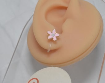 Peut inclure: Une petite boucle d'oreille en forme de fleur rose clair avec une pierre centrale transparente, fixée à un mécanisme à clip en plastique transparent. La boucle est présentée sur un support en forme d'oreille beige avec une étiquette ronde Chrlinge Schmuck ohne Stich.
